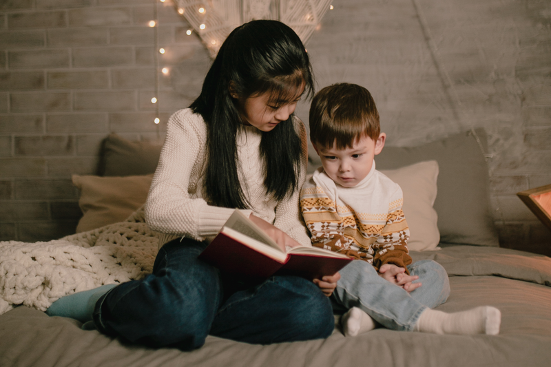 A young girl reads to a little brother on a bed in a room with soft twinkle lights.