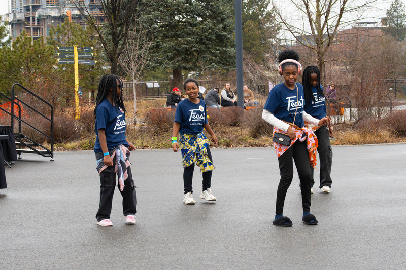 Teenage dancers wearing Feast World Kitchen shirts and coloful scarfs around their waists perform in a park.