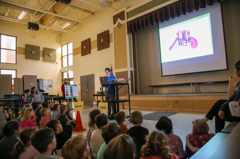 Woman stands in front of a group of children who are looking at a large screen behind her. The screen depicts a slide.
