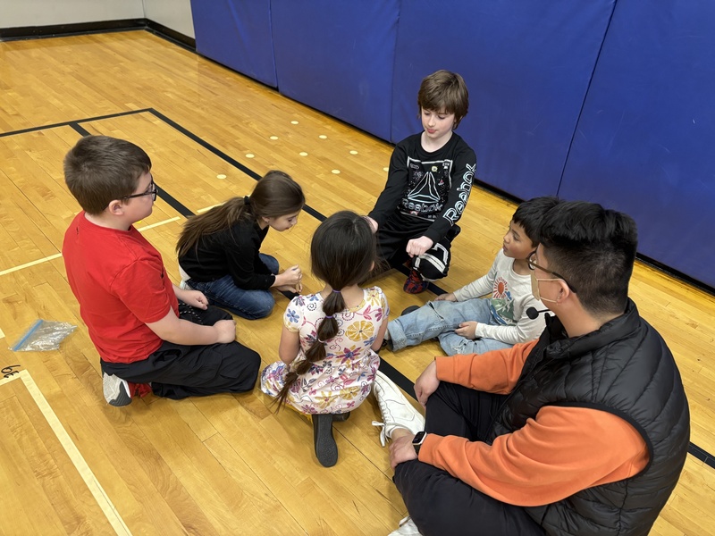 A group of young students sit on the floor of a school gymnasium with a teacher in a cluster.