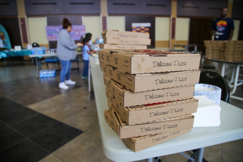 Boxes of pizza sit on a table, with a mother and child in the background.