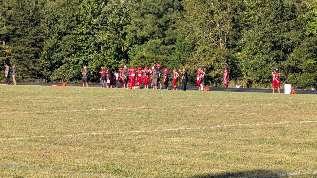 A group of people in red uniforms on a field, standing around a white bucket. Background includes trees.