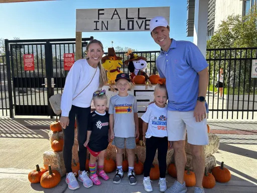 Family standing in front of a pumpkin stand that rads "Fall in Love" on the sign. The stand has stuffed animals of Woodstock and Snoopy.