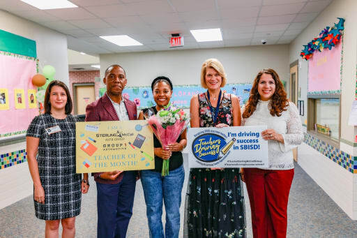 Teacher being presented with a bouquet and signs that say "Teacher of the Month" and "I'm driving success in SBISD" respectively