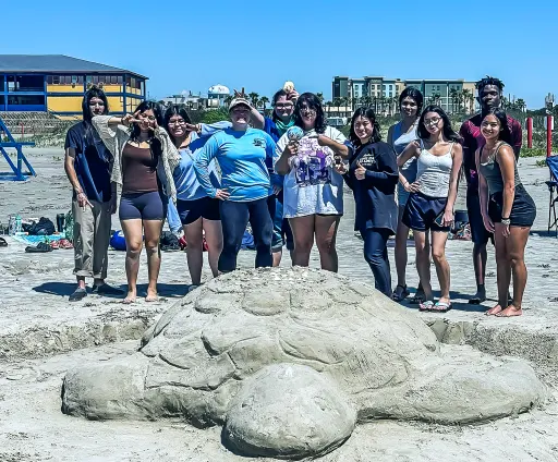 group of high school students grouped behind a sand sculpture of a sea turtle