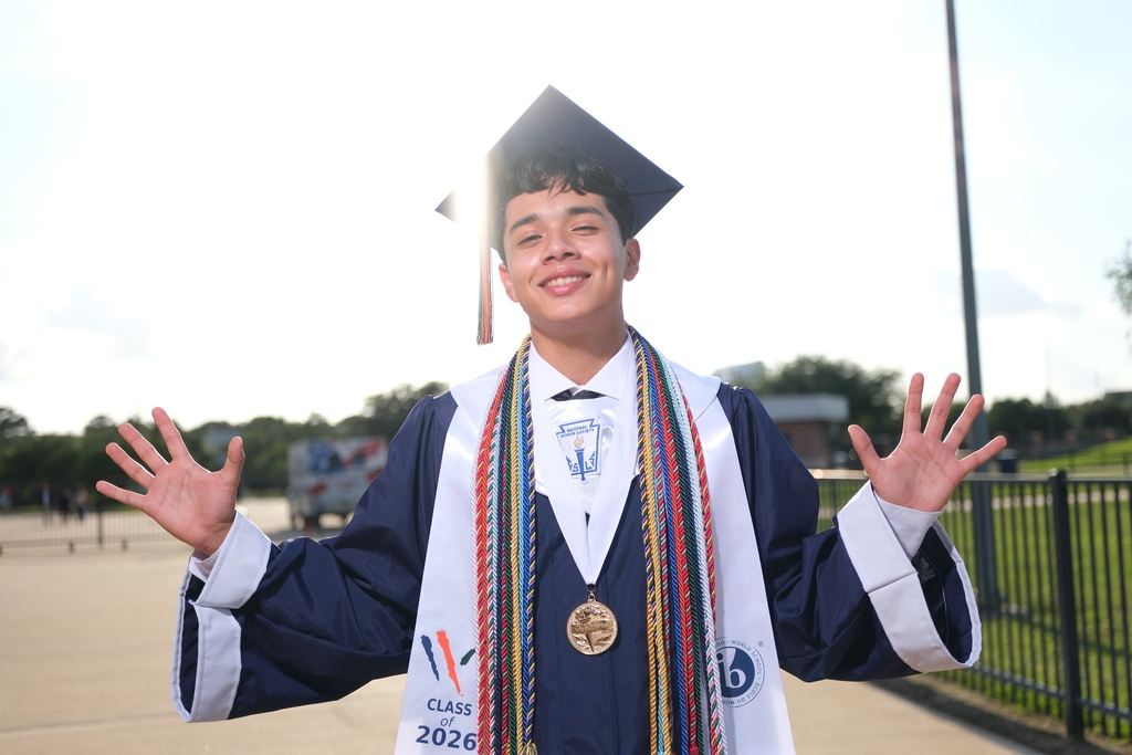 A young man wearing a graduation gown and cap smiles with arms spread wide. A medal hangs on his chest.
