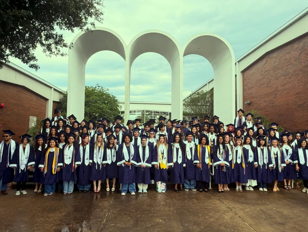 A large group of people in graduation robes stand in front of a brick building with arched structures.