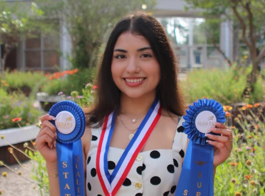 Woman wearing a necklace and a polka-dot dress, holding two blue ribbons and a medal.