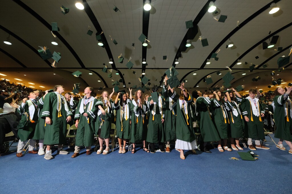 Graduates wearing green gowns and hats celebrate in a hall with blue carpet and lights.
