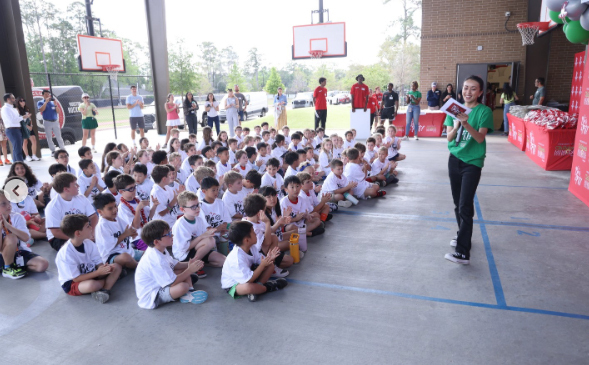 A group of children in white shirts sit on a basketball court with a woman in green.