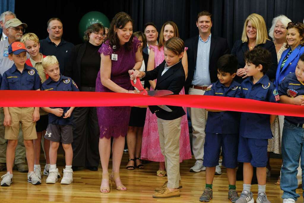 Group of people on stage with red ribbon, two women cutting with scissors, children in uniform.