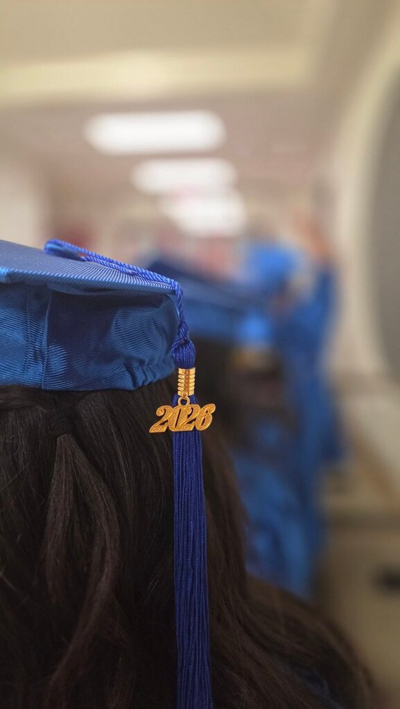 A person wearing a blue graduation cap with a tassel, and a number "2026" is visible. Blurry blue-robed people in the background.