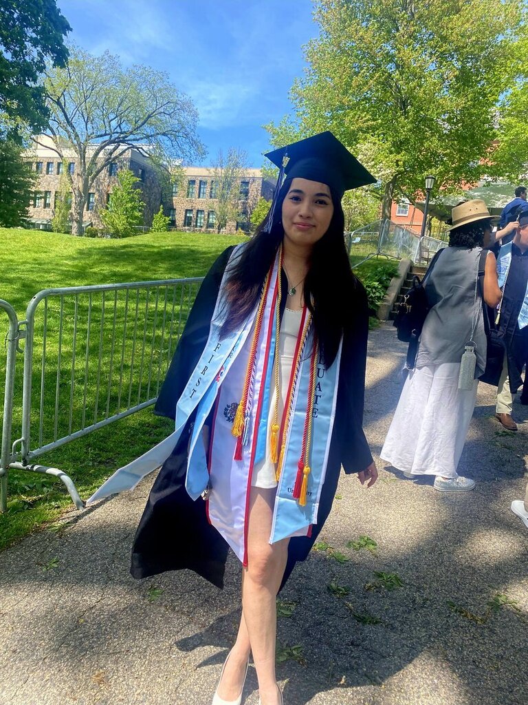 A woman in a graduation gown stands on a path, surrounded by a fence and greenery.