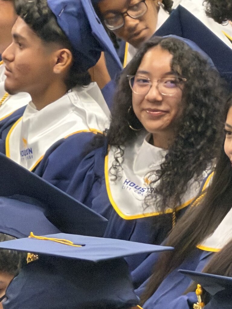 Graduates in caps and gowns, some wearing glasses, seated in an auditorium with words "HOUSTON" on their robes.