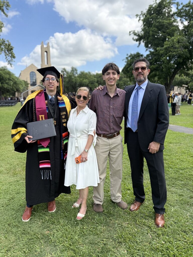 Four people stand on green grass. One holds a diploma. A church is behind them.
