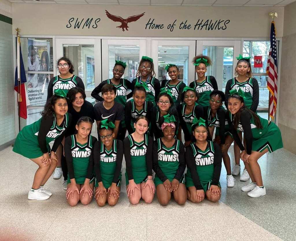 Cheerleaders in green uniforms pose for a group photo in front of a sign reading "Home of the Hawks".