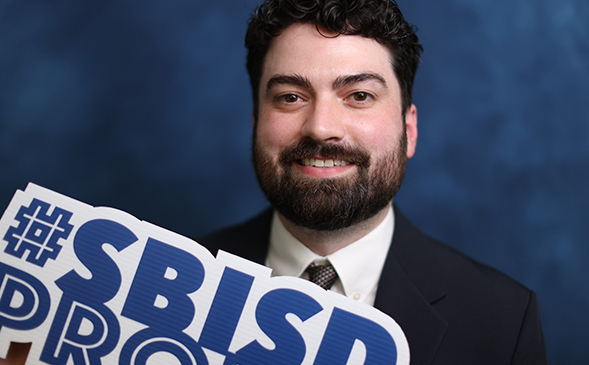 Man with a beard and dark suit, smiling while holding a white sign with blue text that reads "SBISD PRO".