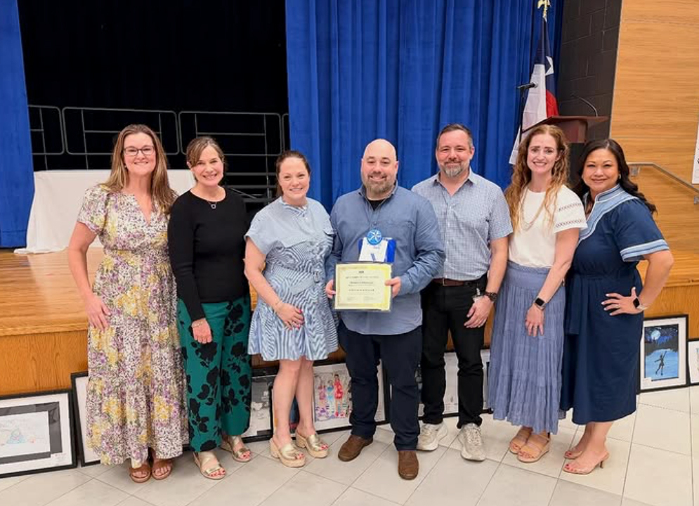 A group of six adults in dresses or blouses and slacks stand together, one holding a plaque. A stage with blue curtains is behind them.