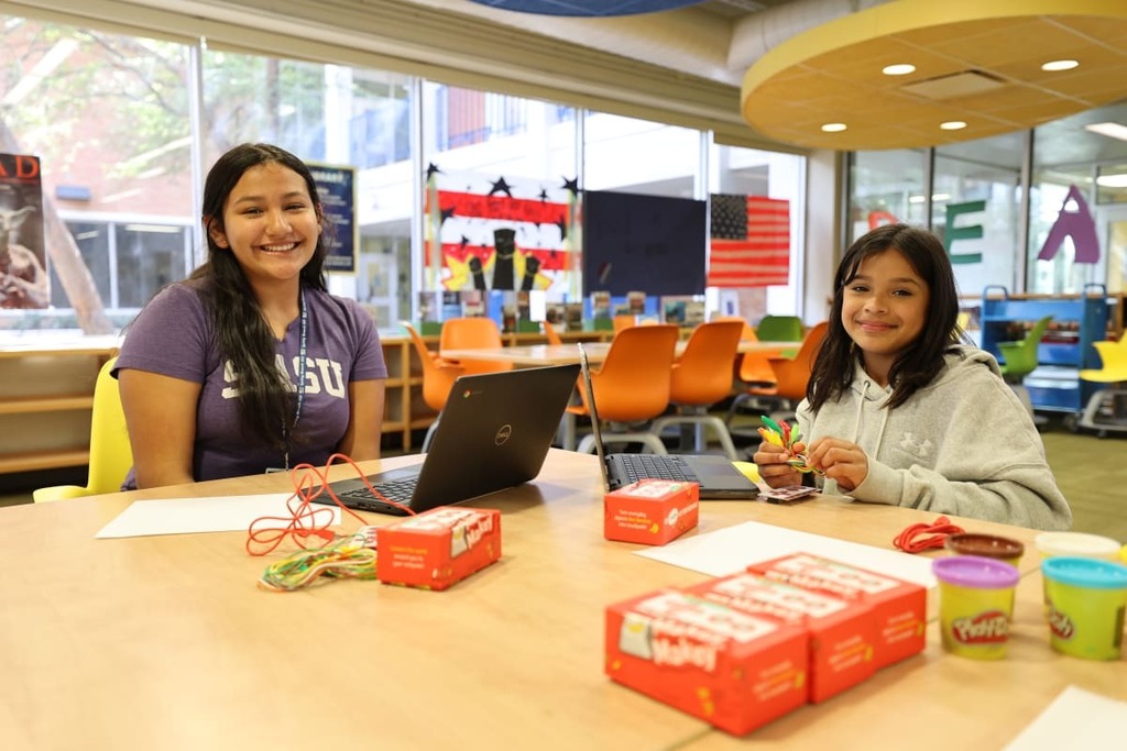 Two young women seated at a table with laptops, a box of craft supplies, and colored cups.