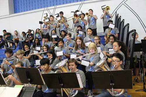 A group of people in blue shirts playing instruments in a room with bleachers and a blue wall.