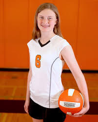 Girl in white jersey with number 6 stands in a gym, holding a volleyball. Background is an orange wall.