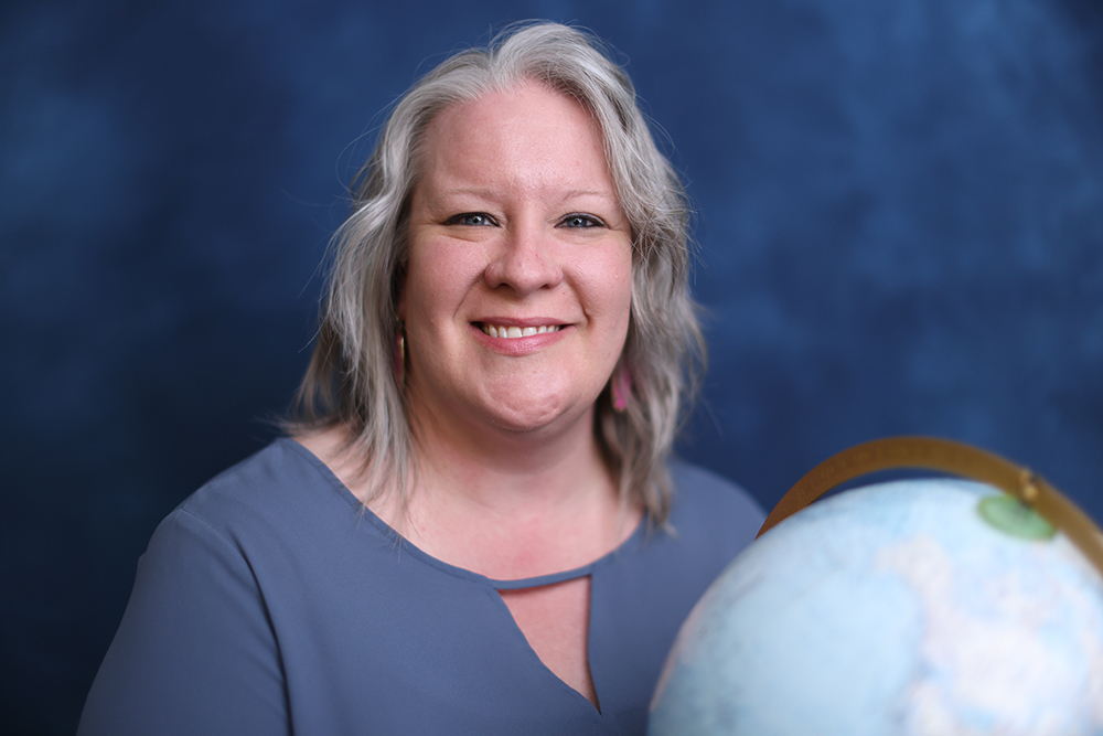A smiling woman with gray hair holds a globe against a blue background. She wears a blue blouse.