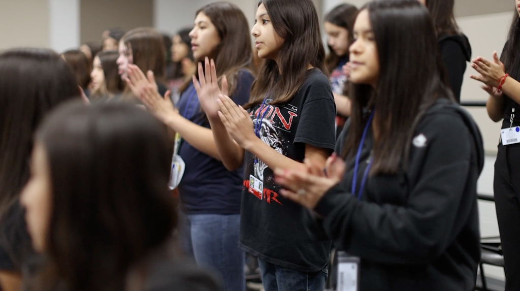 A group of young women wearing black tops and jeans stand in a room, clapping hands.
