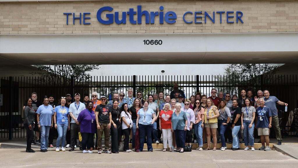 A group of people stands in front of a building. The sign reads "The Guthrie Center 10660."