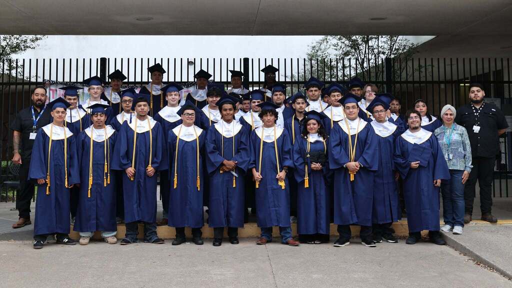 A group in blue gowns and caps, possibly for a graduation, standing in rows in front of a fence.