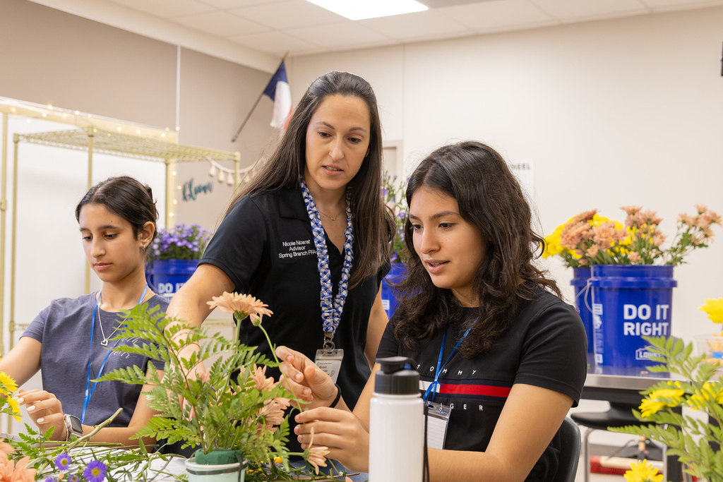 Three people in a classroom setting, two students arranging flowers and one teacher assisting.