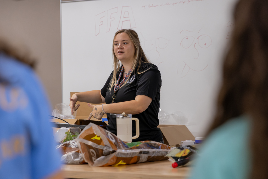 Woman stands at a table, facing a whiteboard with red writing, with a small group of people watching.