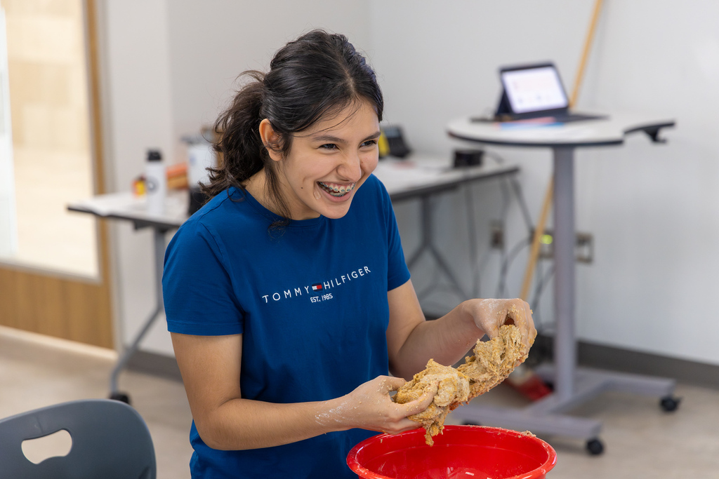 A woman smiles while holding dough in a room with a desk, a laptop, and a chair.