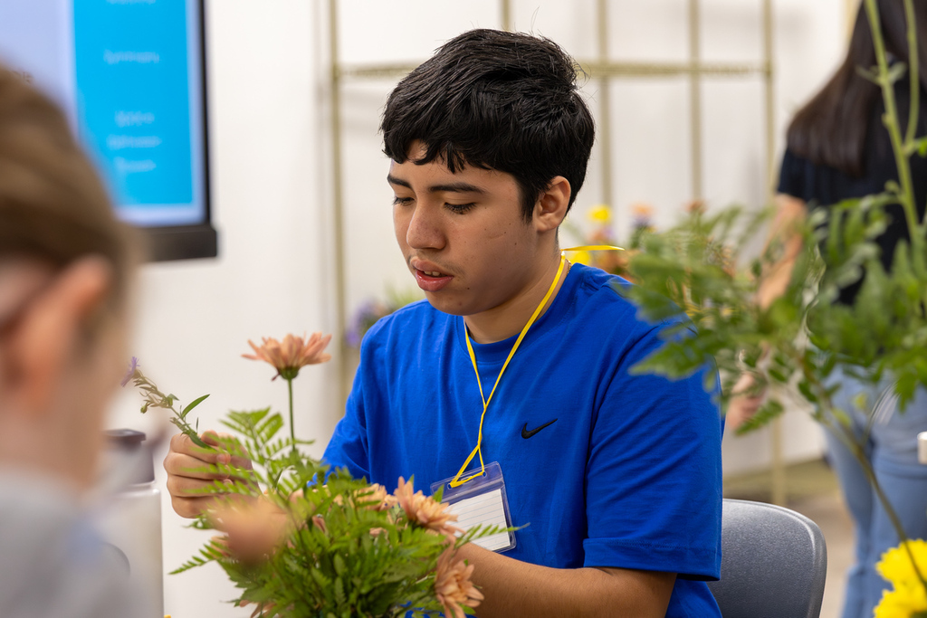 A man in a blue shirt is arranging flowers with others in a room. A monitor is visible in the background.