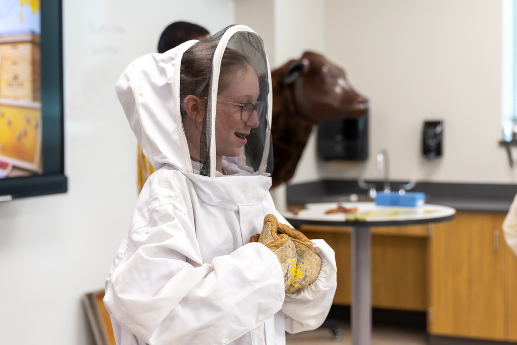 A person in a beekeeper's suit stands next to a cow head and a table with various items.