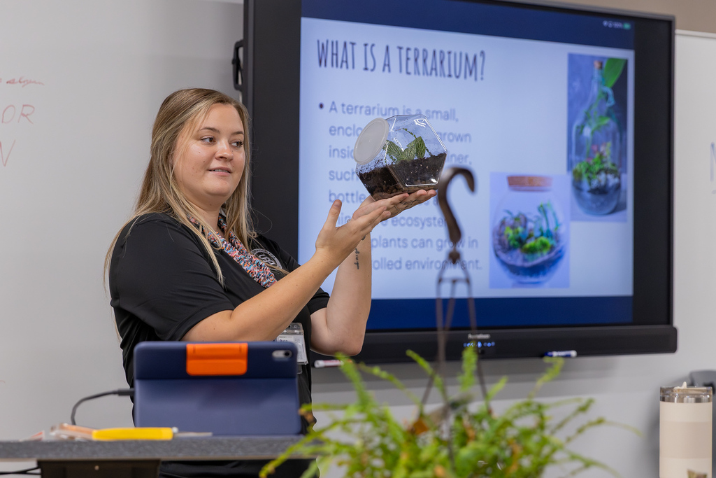 Woman in black shirt holding a small plant in pot at front of classroom with screen projector.