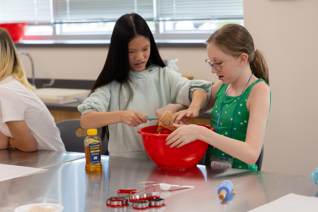 Two girls in a kitchen, mixing dough in a red bowl. A bottle of oil and other tools are on the table.