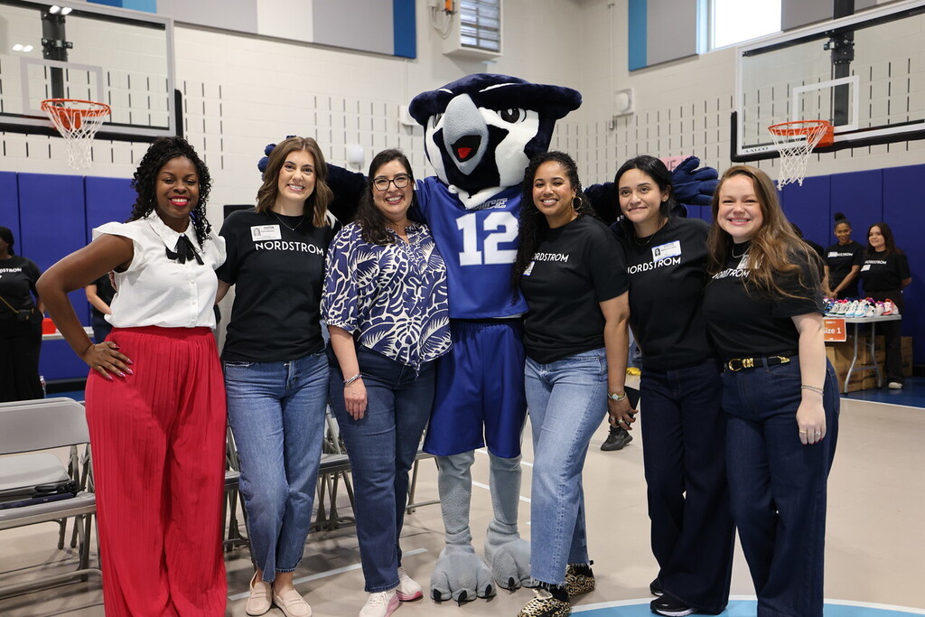 Seven women in matching shirts, one in a sports mascot costume, pose in a gym.