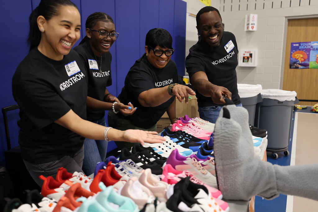 A group of four people in black shirts stand around a table with baby shoes. One person points to a shoe.