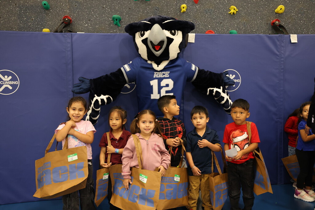 Several children stand in a group with a mascot in a gym. The mascot wears a sports jersey.