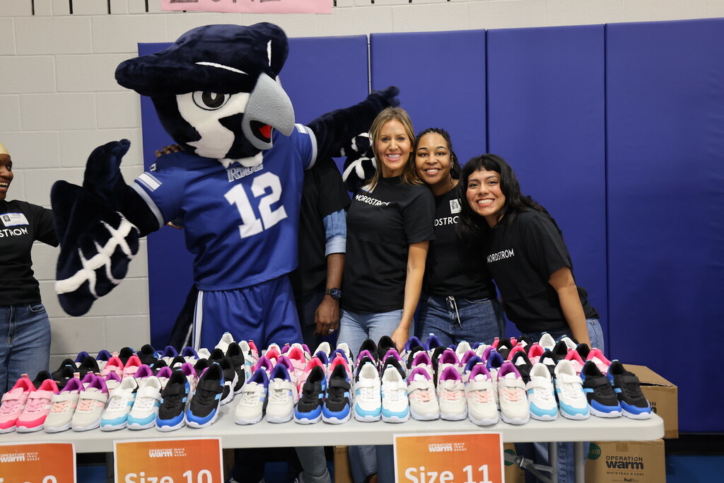 A group of women pose with a mascot and a table of shoes, sizes 10 and 11.