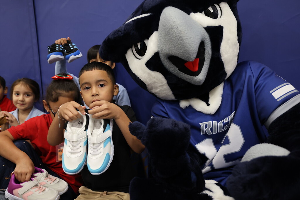 A young boy holds two white shoes in front of a mascot, with children behind him. The mascot wears a blue jersey.