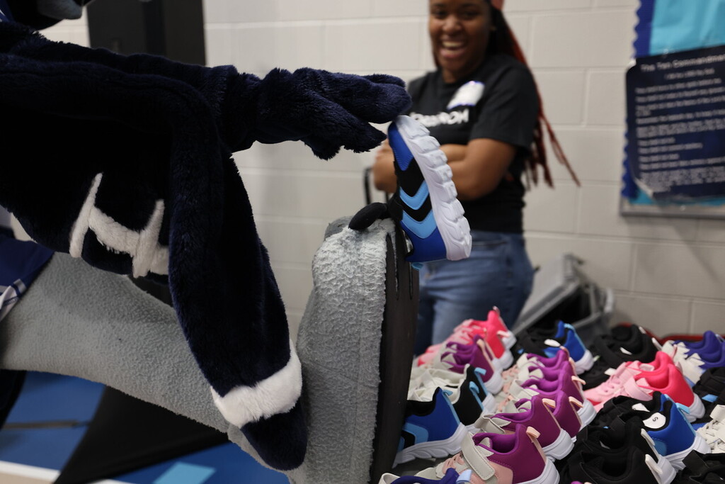 A person holds a shoe while a woman in a black shirt and jeans stands behind them. Colorful sneakers are on a table.