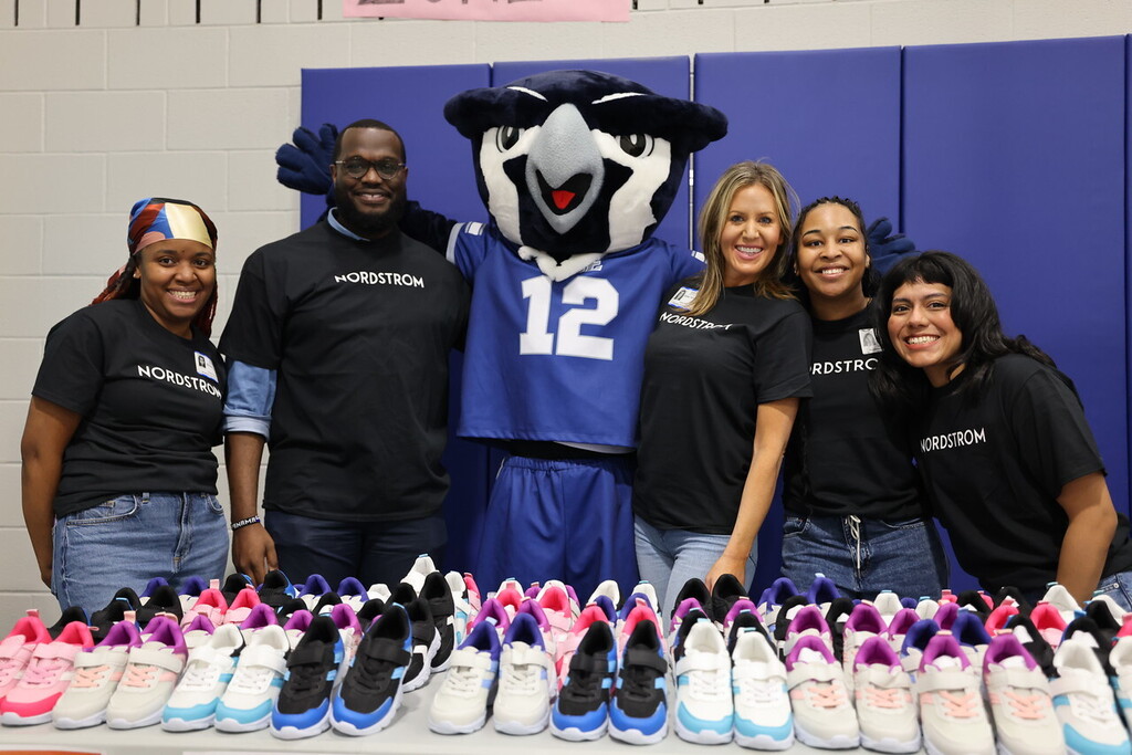 A group of people in matching black t-shirts stand behind a table of sneakers with a mascot.