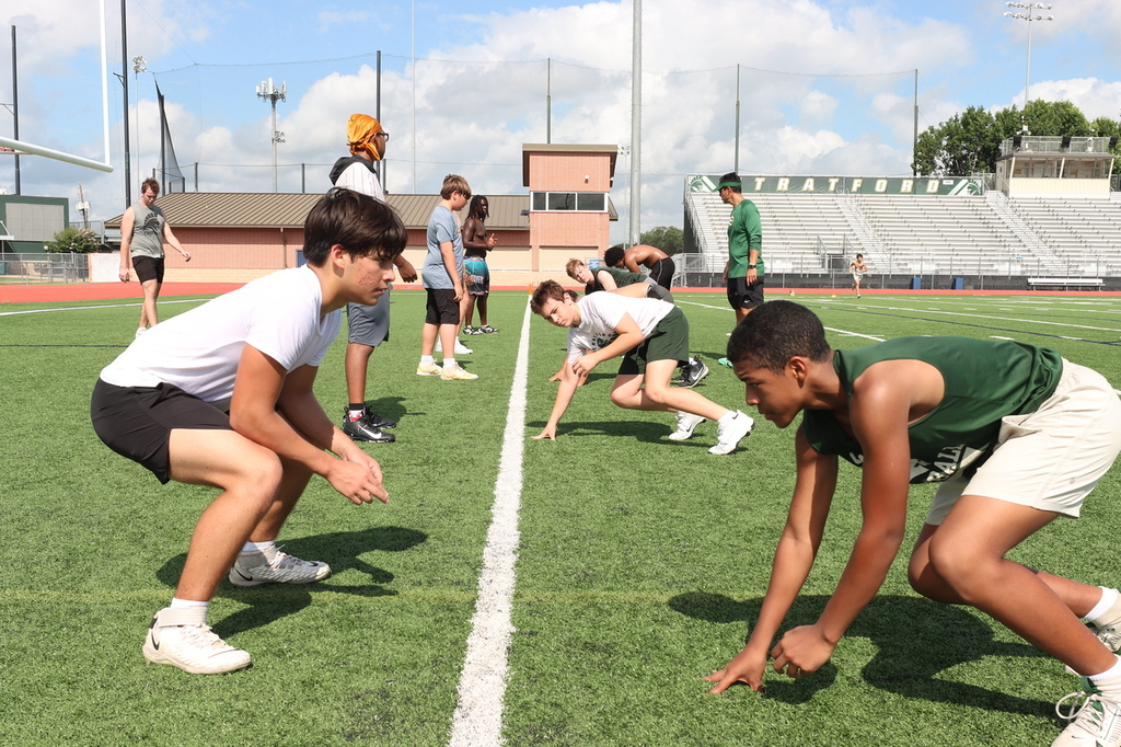 A group of people in sports attire doing exercises on a field, with bleachers and a building in the background.