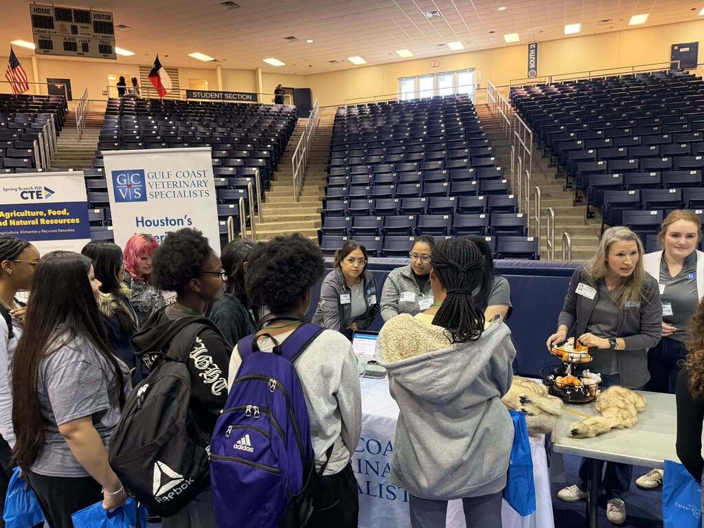 A group of people gather around tables in an auditorium, engaging in conversation. Tables have food and promotional materials.