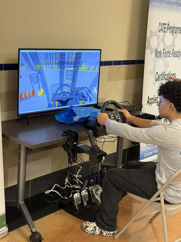 Person seated at a desk, interacting with a computer monitor and steering wheel, in a room with a banner.