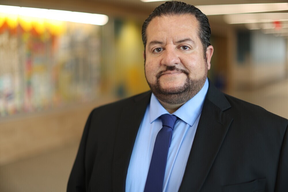 A man with stands in a hallway, wearing a black suit, a blue shirt, and a blue tie.