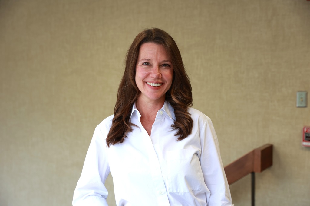 Woman with brown hair stands near a staircase, wearing a white shirt and smiling at the camera.
