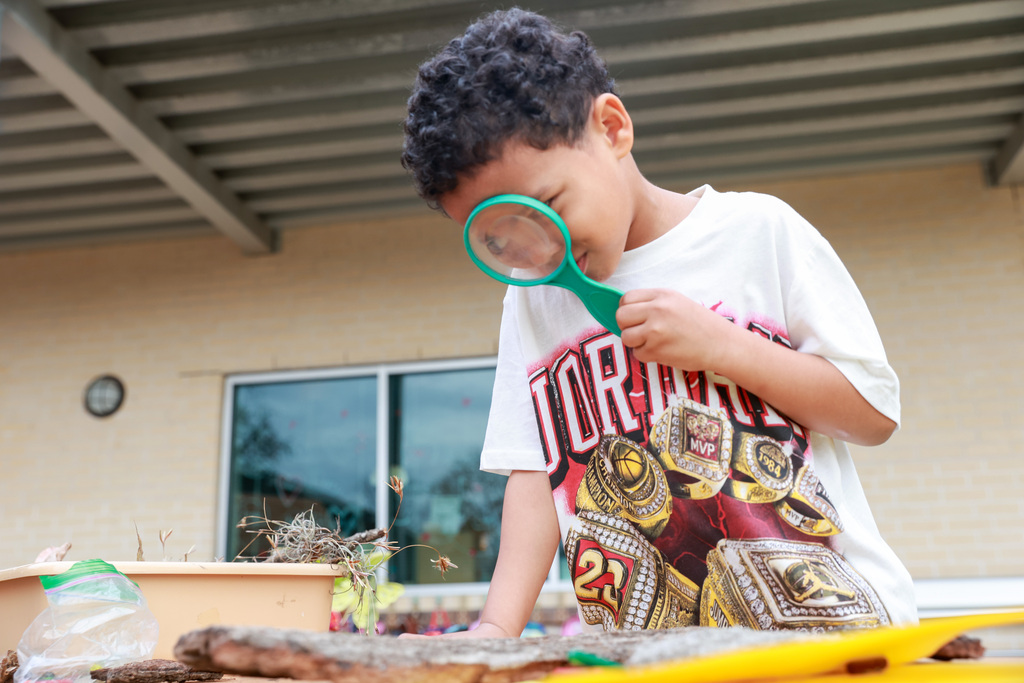 A young child examines a rock using a magnifying glass. Behind them, a brick wall and a glass door.