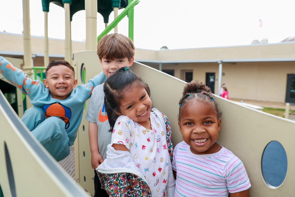 Four children on a playground, one standing, two on a slide, and one climbing, all smiling.